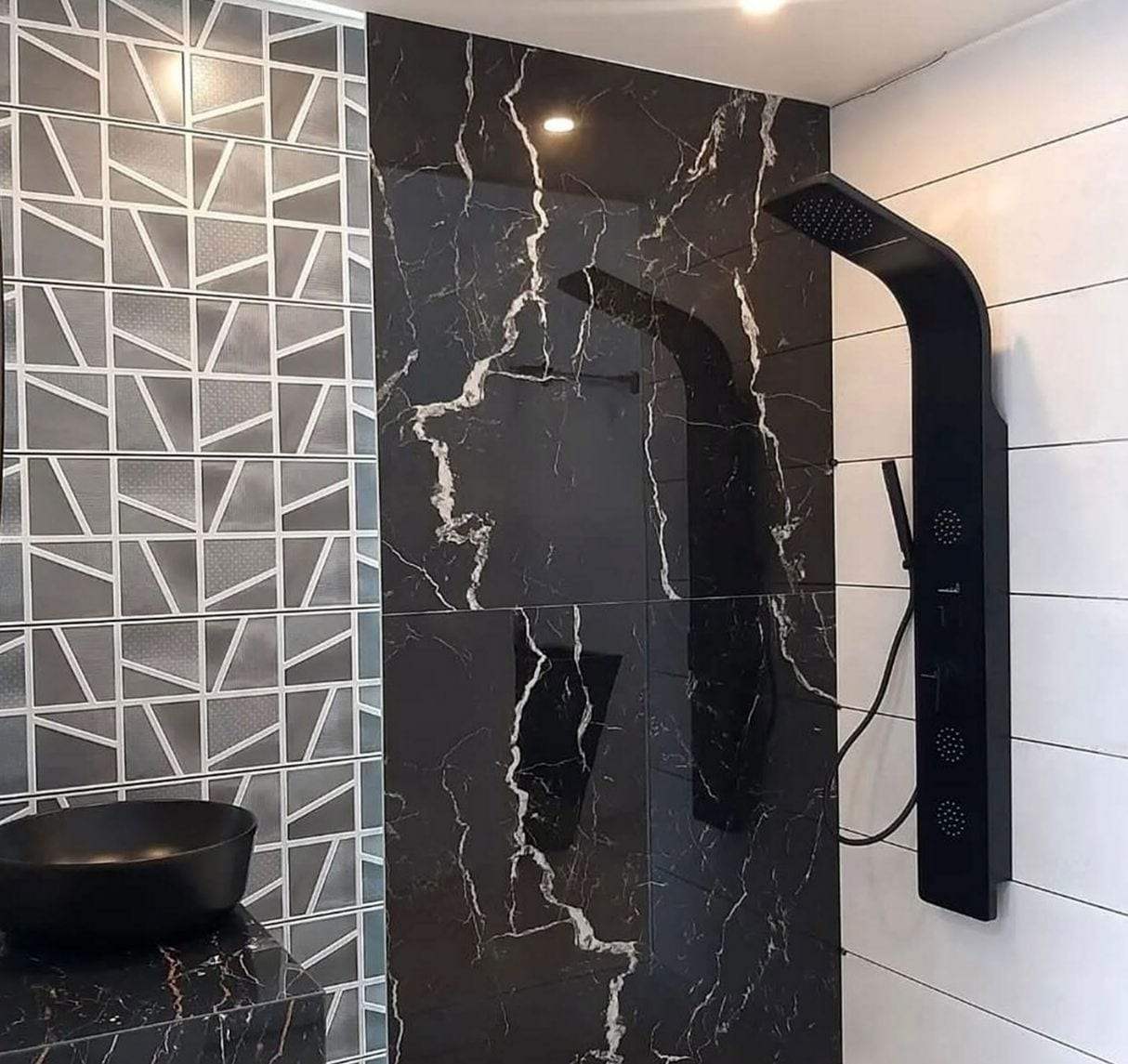Matte black shower panel with a rain head, body jets and a hand shower mounted on black marble-look tile, beside a black vessel sink on a marble counter