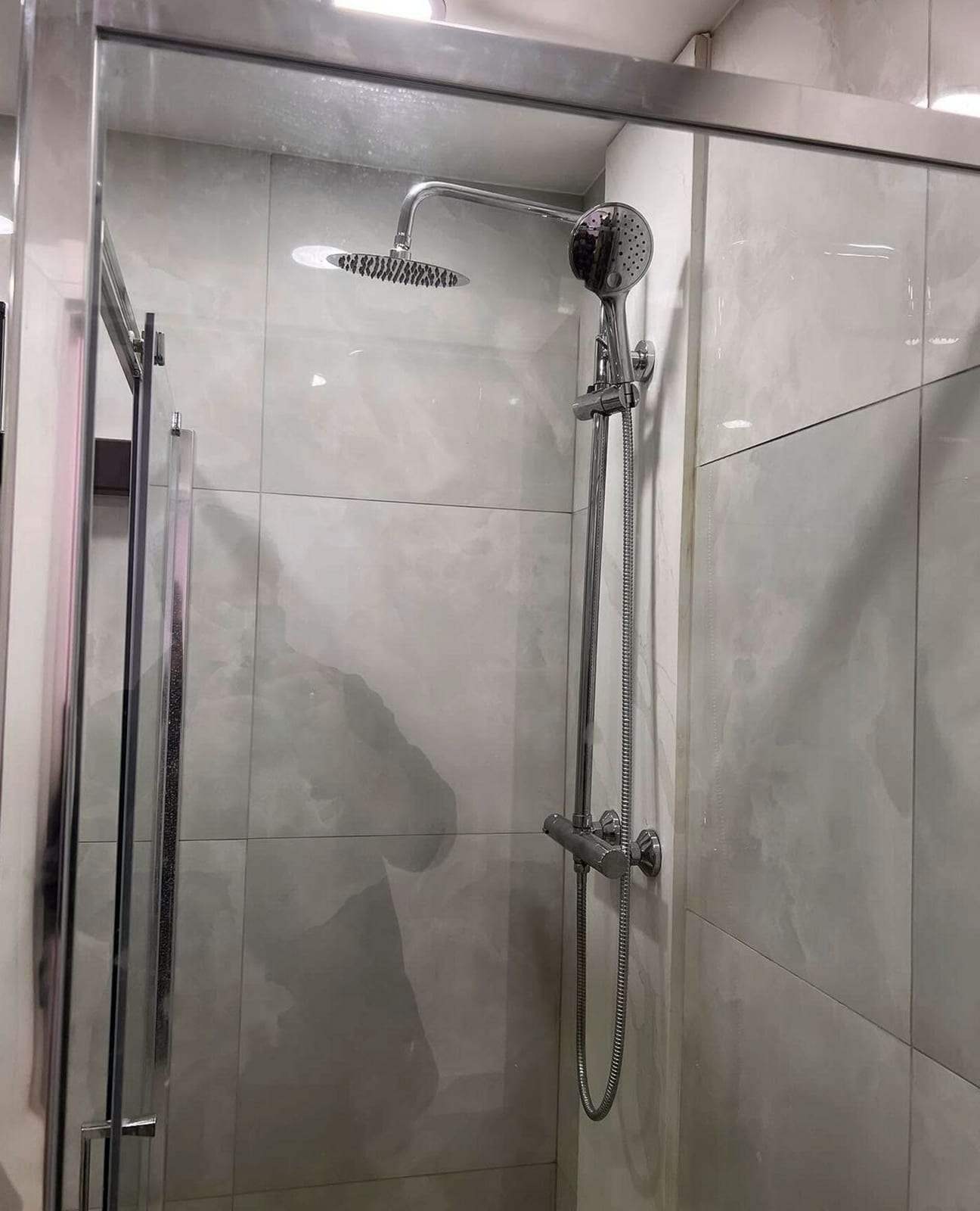 Glass shower enclosure with a chrome thermostatic shower bar, a round rain head and a hand shower on a slide rail, against light gray marble-look tile