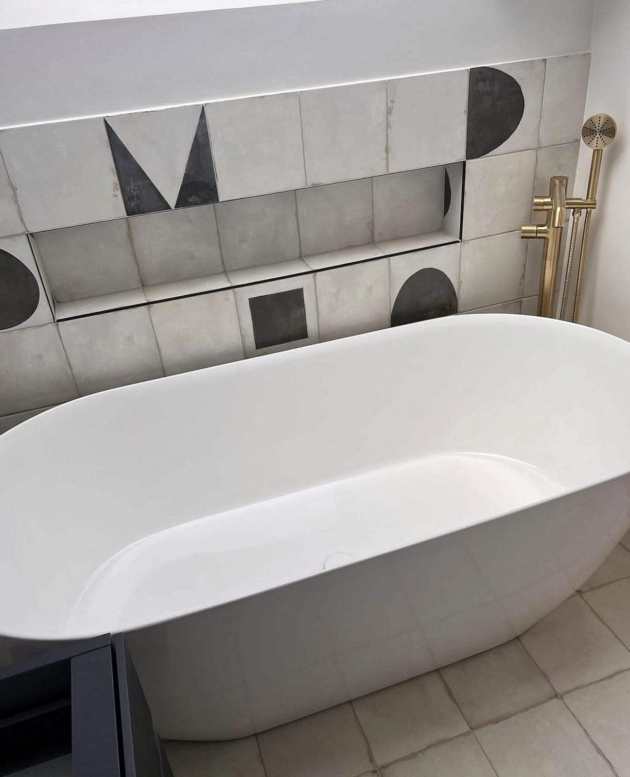 White freestanding soaking tub in front of patterned tile with a recessed niche, with a brushed brass floor-mounted tub filler and hand shower at one end