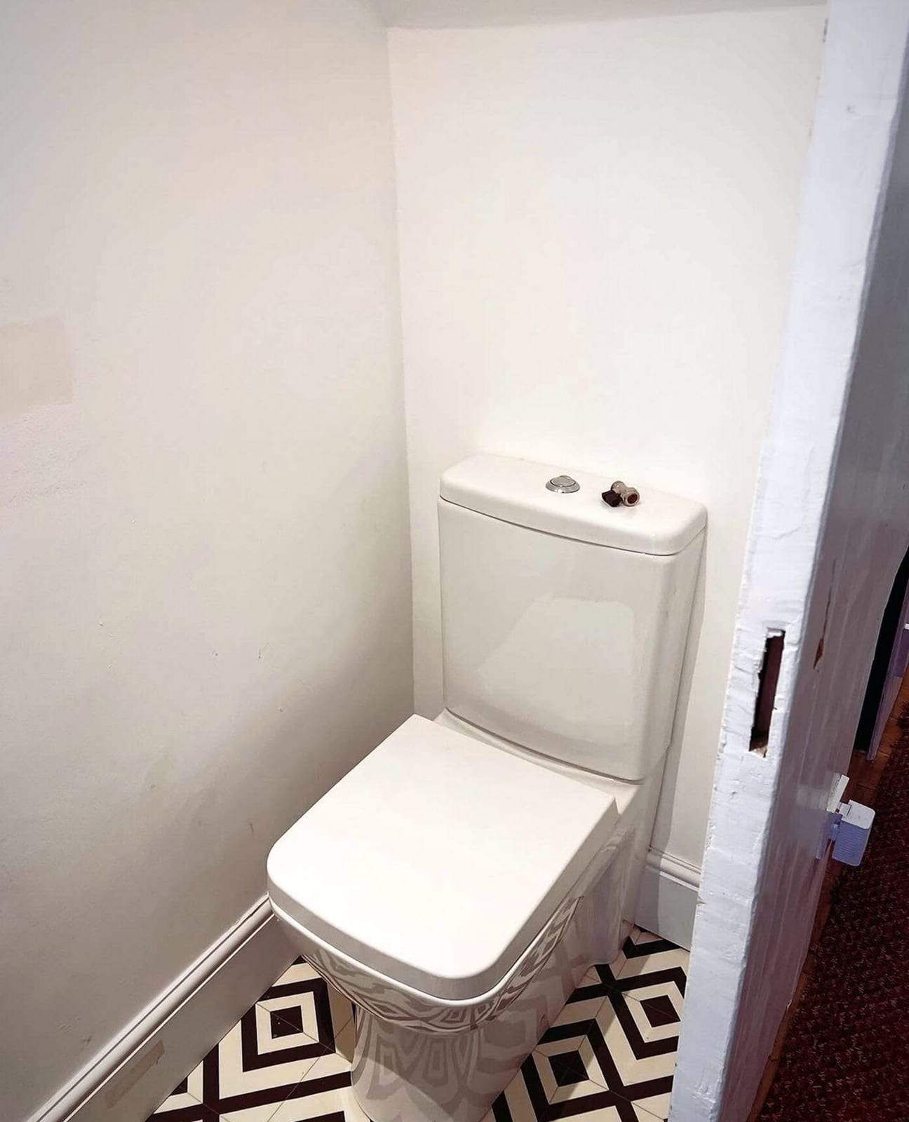 Newly installed white close-coupled toilet with a square seat in a narrow half bath with plain white walls and black-and-white patterned floor tile