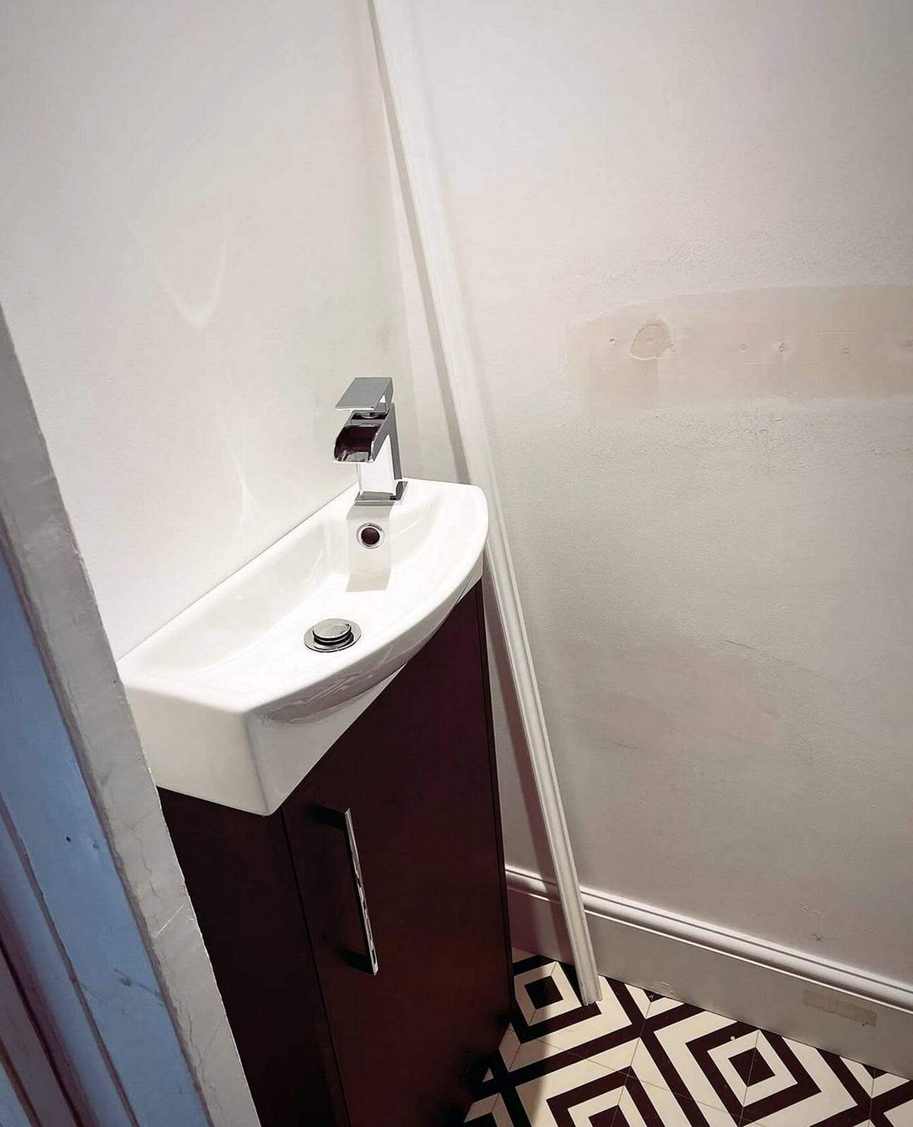 Compact white vanity basin with a chrome waterfall faucet on a dark wood cabinet in the corner of a half bath with patterned floor tile