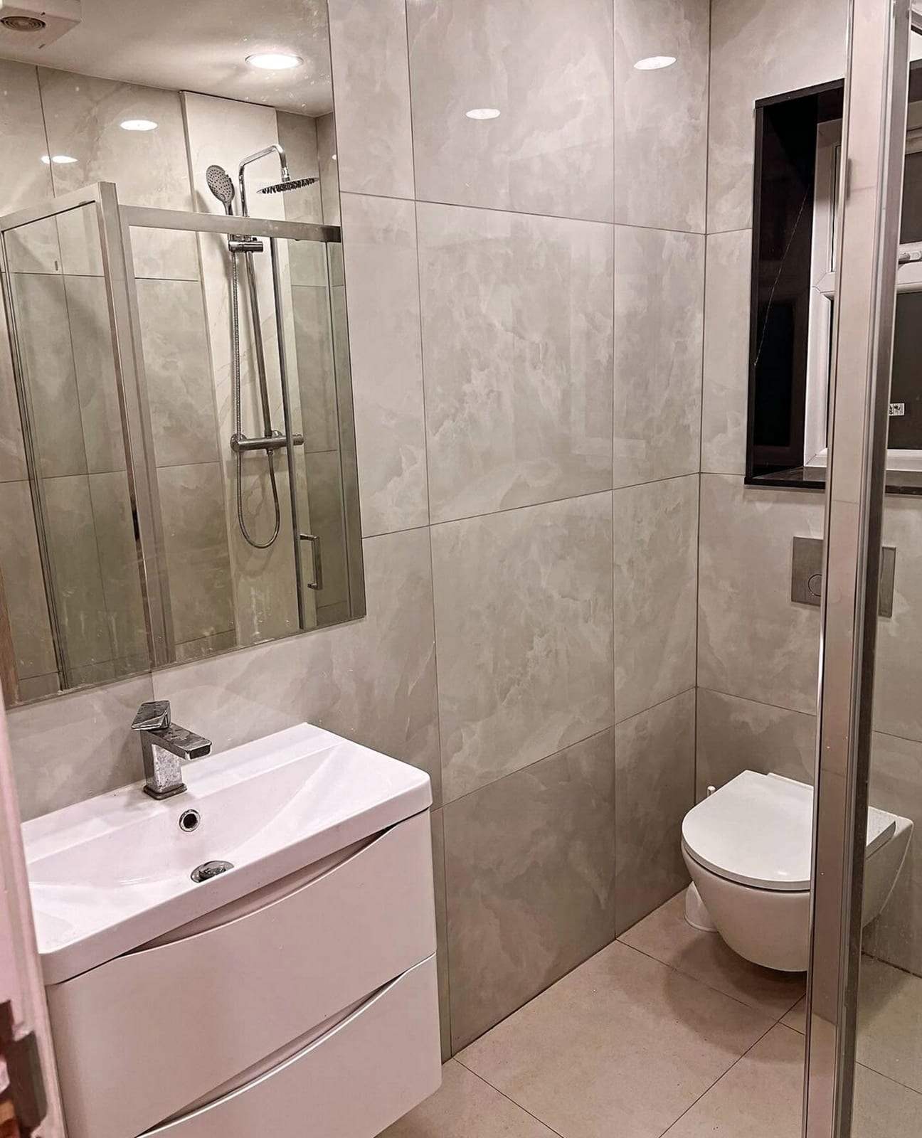 Finished bathroom in light gray marble-look tile with a white wall-hung vanity and chrome faucet, a wall-hung toilet with a wall flush plate, and a shower enclosure reflected in the mirror
