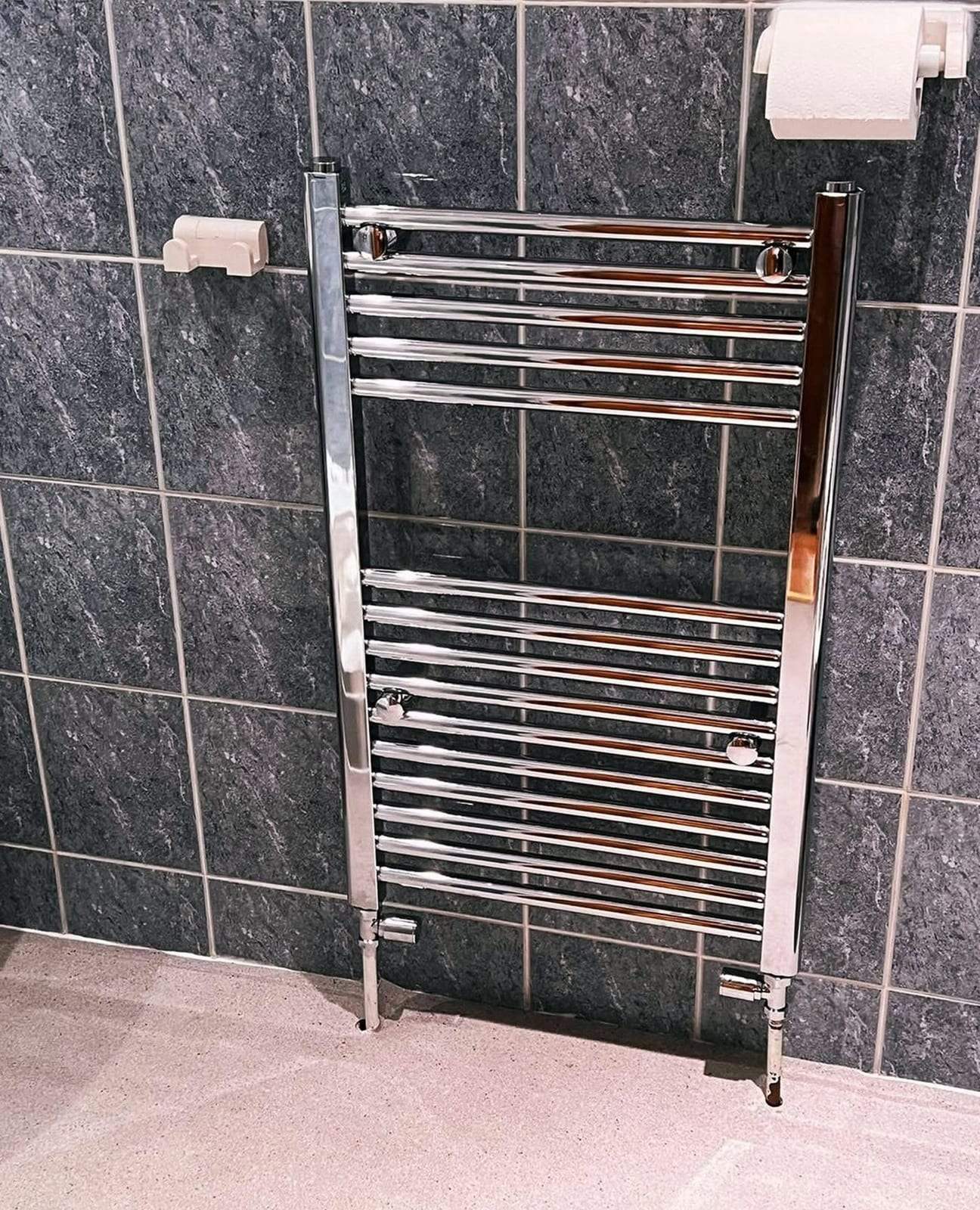 Chrome ladder-style towel warmer connected to two chrome valves at floor level, mounted on dark slate-look wall tile