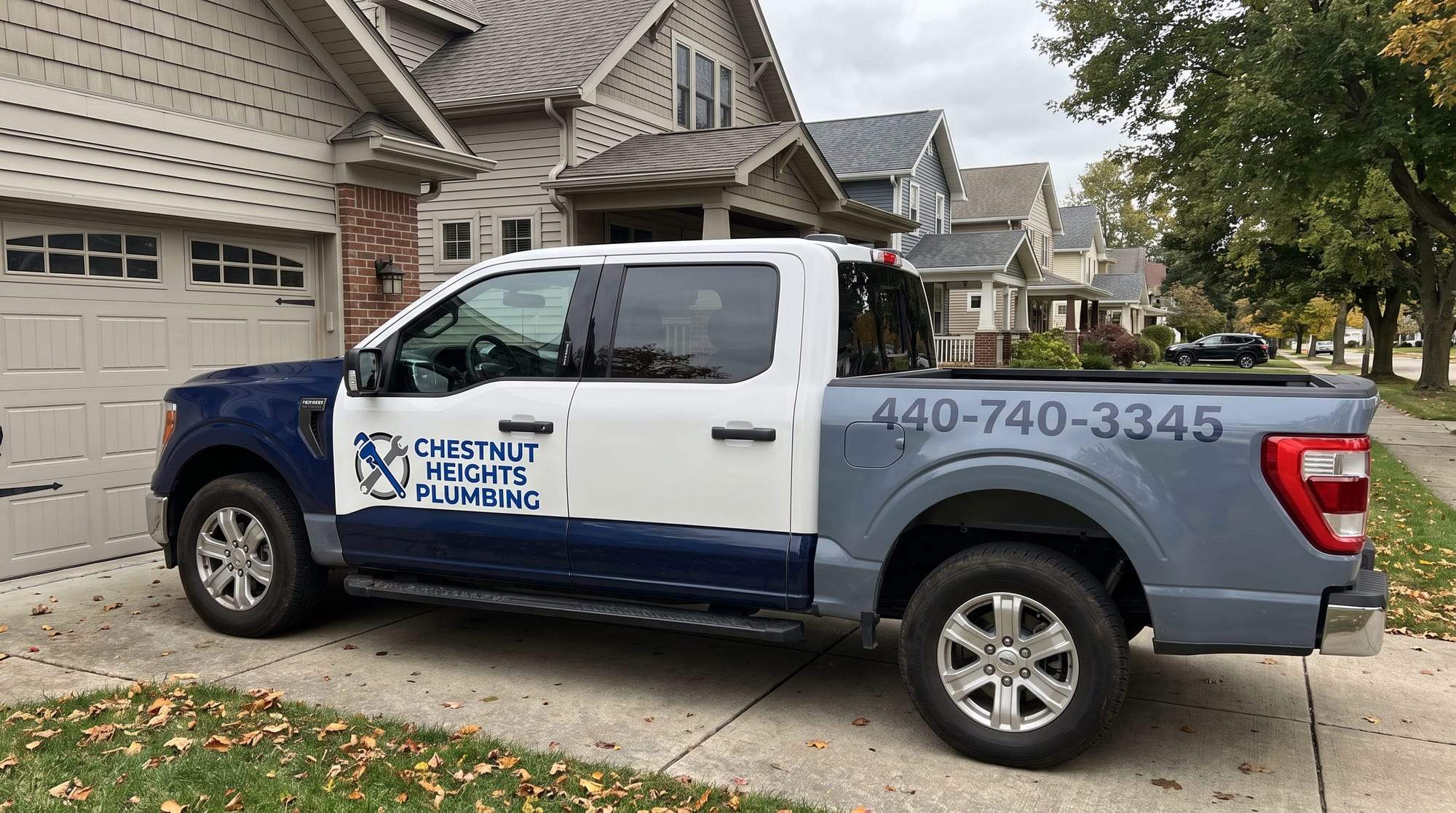 White Chestnut Heights Plumbing pickup truck with navy lower panels, the company logo on the door and 440-740-3345 on the slate-gray bed, parked on a concrete driveway on a tree-lined residential street in autumn