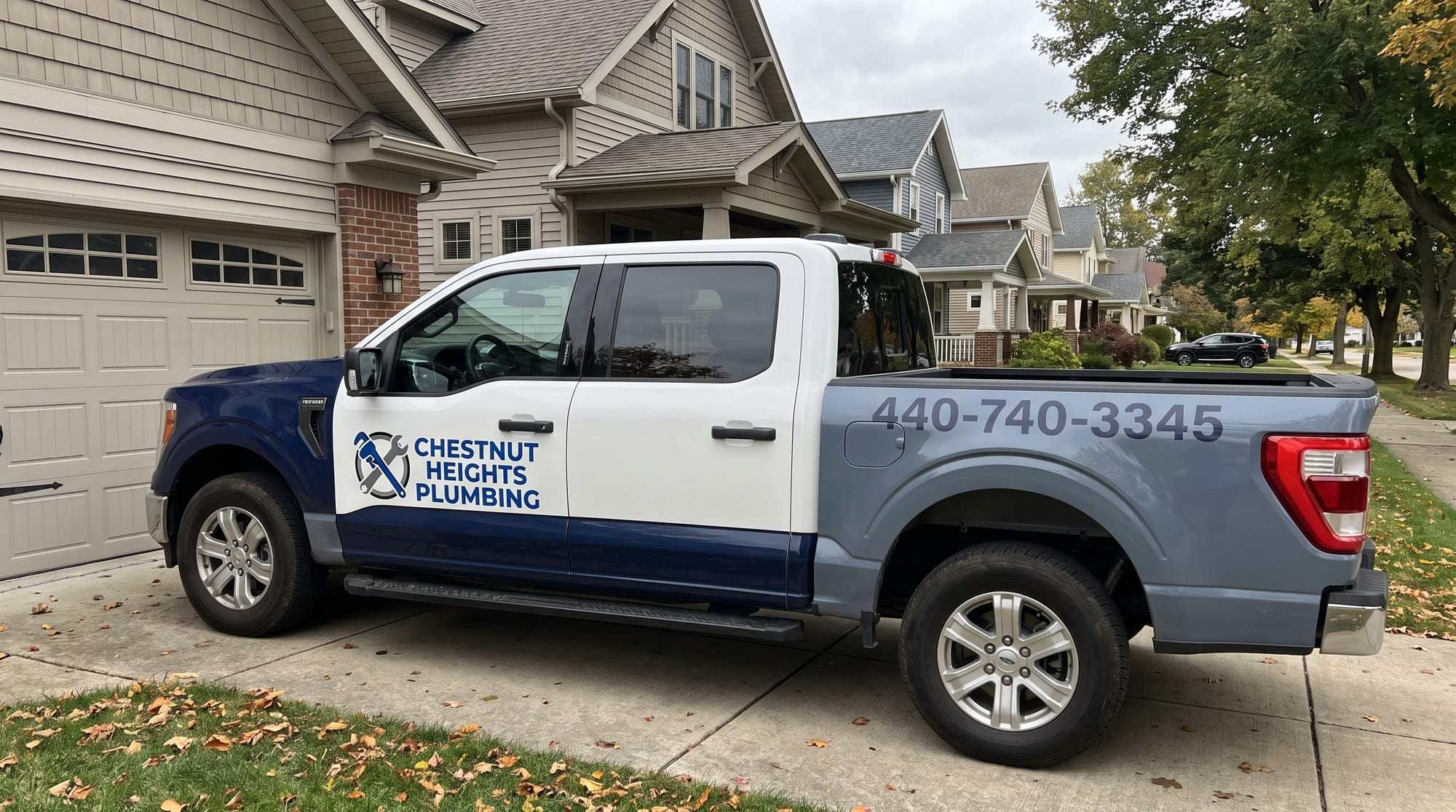 White Chestnut Heights Plumbing pickup truck with navy lower panels, the company logo on the door and 440-740-3345 on the slate-gray bed, parked on a concrete driveway on a tree-lined residential street in autumn